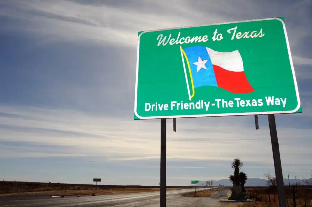 A green road sign that says, “Welcome to Texas. Drive friendly—The Texas Way” marks the Texas state line on a remote country road.