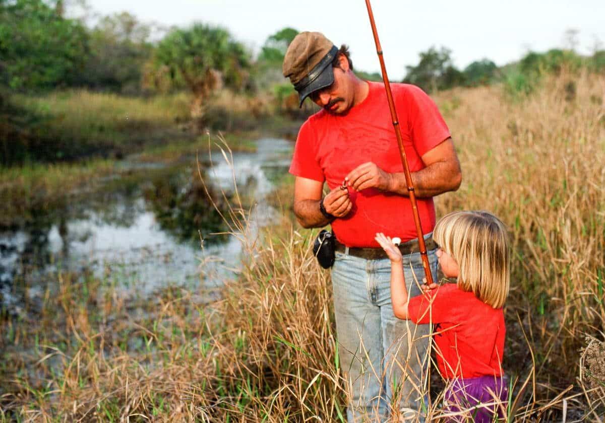 A father baits the hook while the daughter holds the fishing rod beside a pond.