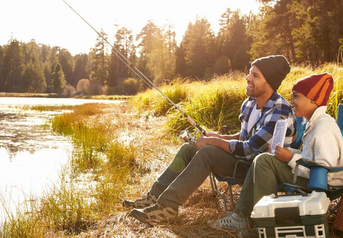 A smiling father and son fish by a river during a fall sunny day.