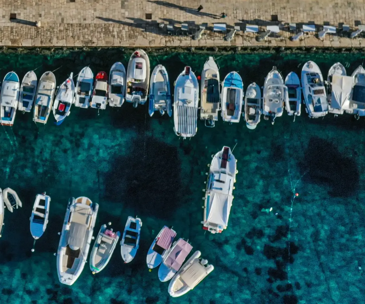 Aerial view of numerous small boats and yachts docked along a stone pier in clear turquoise water.