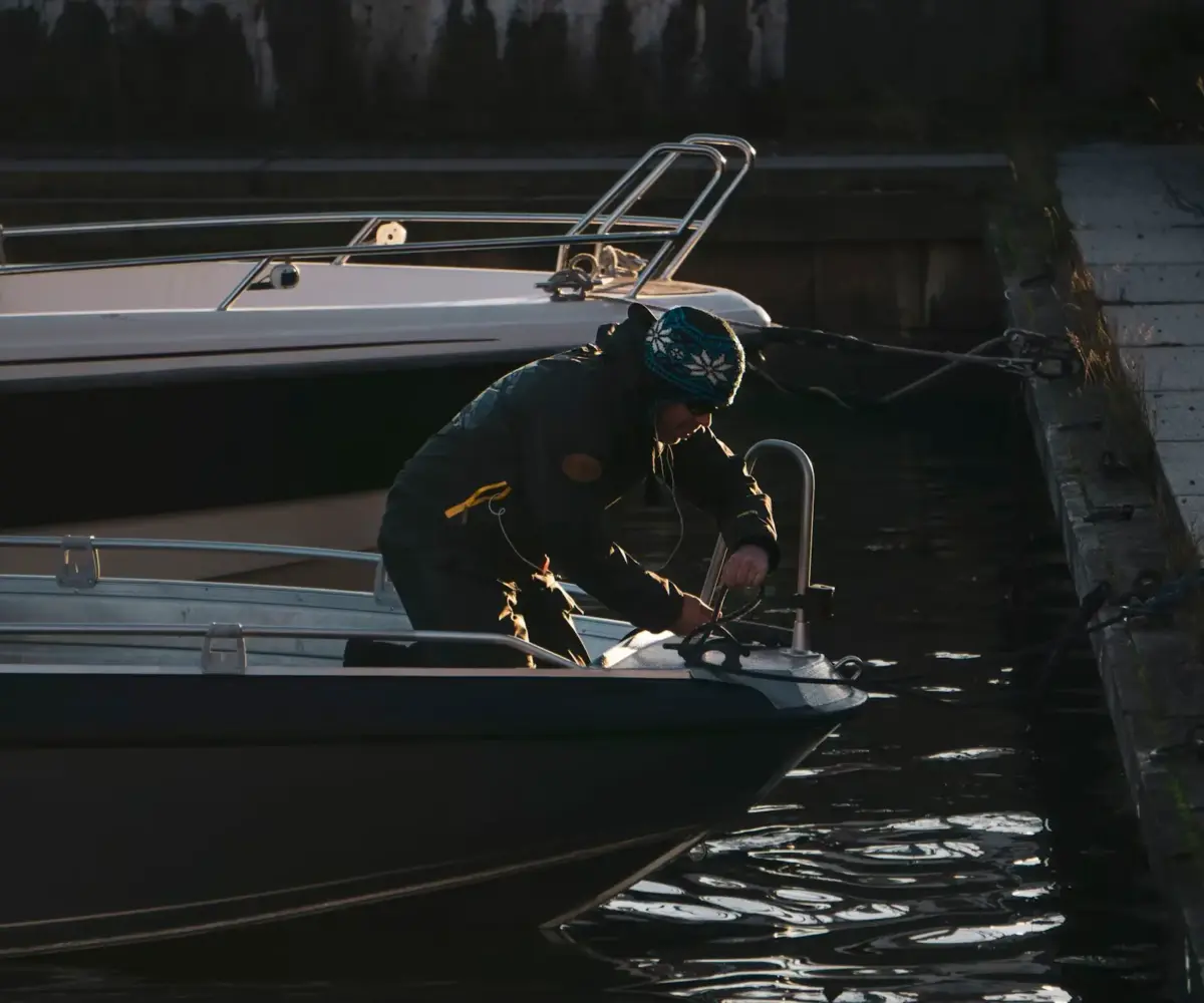 Person maintaining and securing a small motorboat to a dock in low evening light.