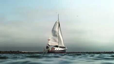 A sailboat with fully raised sails gliding through calm waters near a rocky breakwater under a cloudy sky.