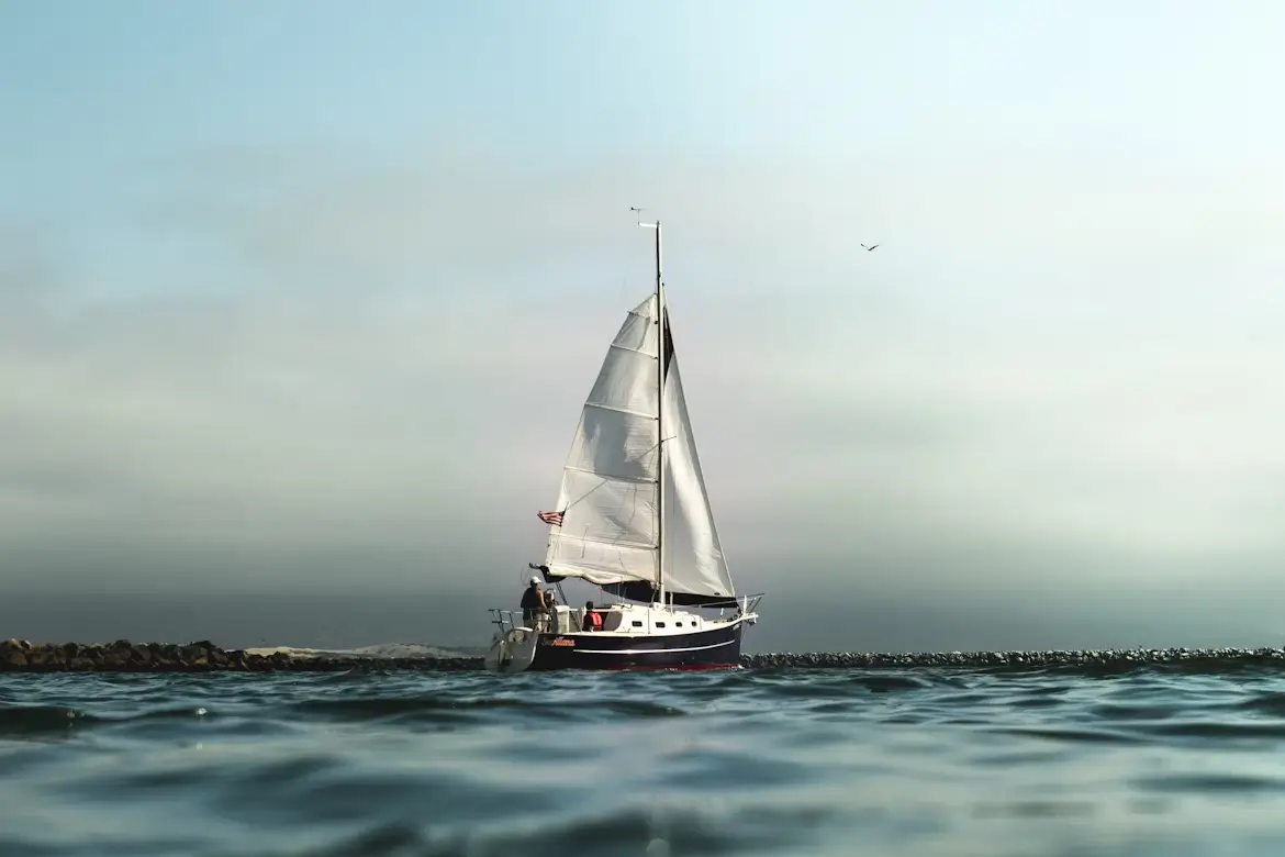 A sailboat with fully raised sails gliding through calm waters near a rocky breakwater under a cloudy sky.