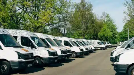 A row of white commercial vans parked in a lot surrounded by trees on a clear day.