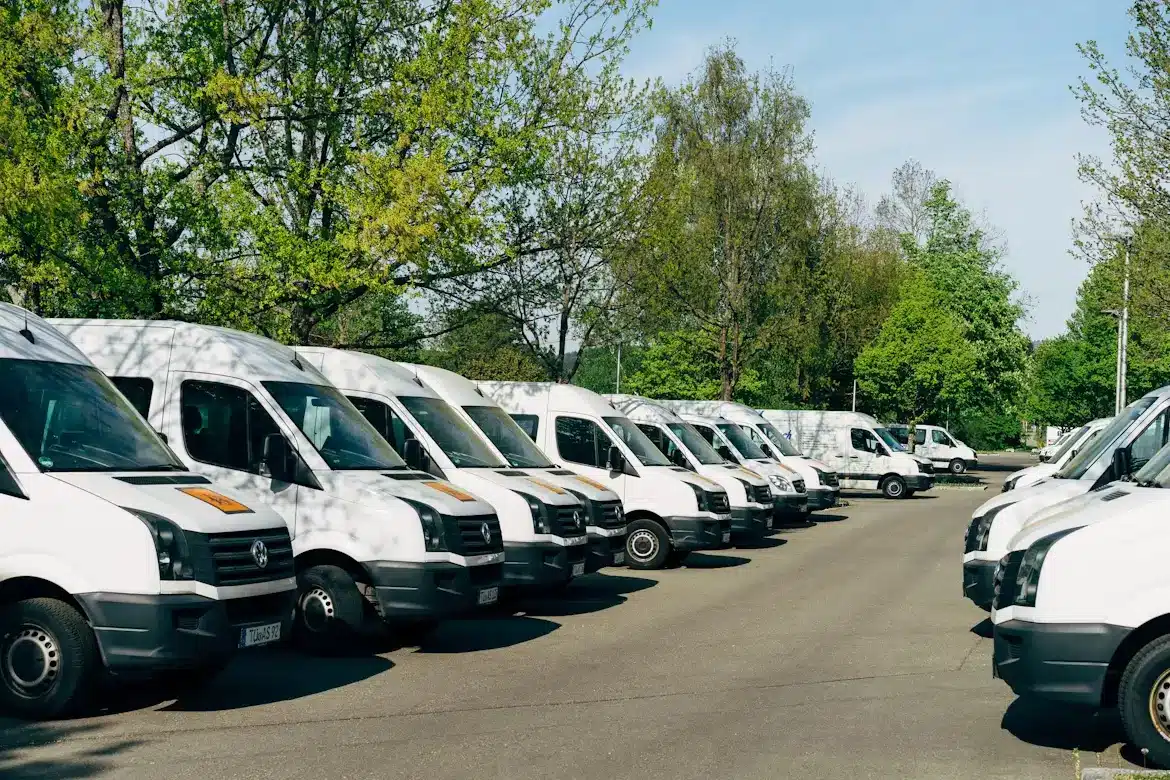 A row of white commercial vans parked in a lot surrounded by trees on a clear day.
