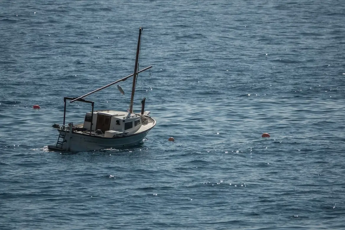 A small fishing boat anchored alone in calm, open blue water with a few floating buoys nearby.