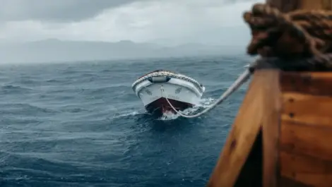 Small boat being towed through rough ocean waters on a cloudy day.
