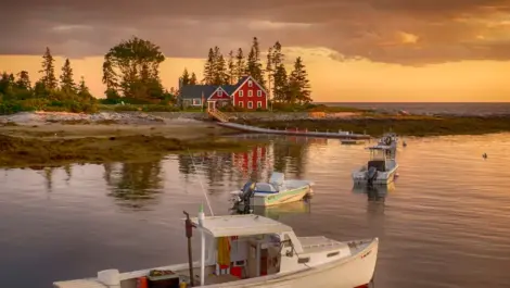 Small boats anchored near a coastal home with a red exterior, surrounded by trees, during a golden sunset.