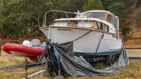 Covered boat on a trailer stored in a grassy yard with a red inflatable boat beside it.