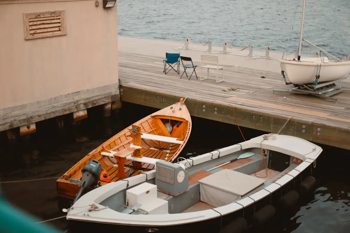 Two small boats tied at a wooden dock with chairs and a sailboat nearby.