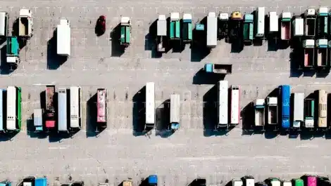 Aerial view of a large truck parking lot with multiple rows of parked semi-trucks, trailers, and a few smaller vehicles.