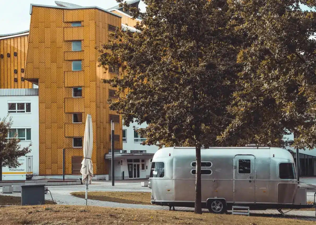 A silver trailer parked under a tree near a yellow modern building.
