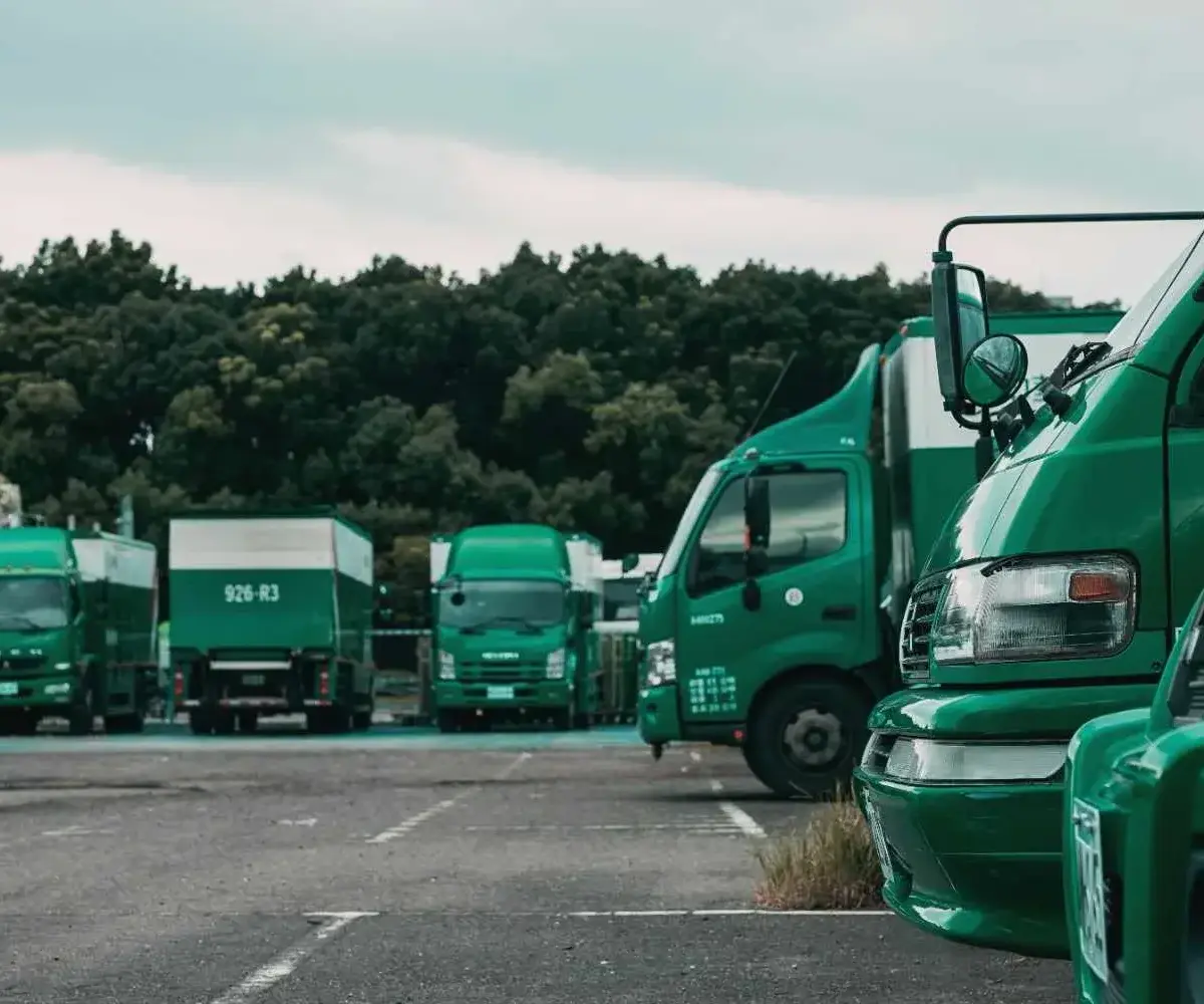 Green delivery trucks parked in a lot with trees in the background.