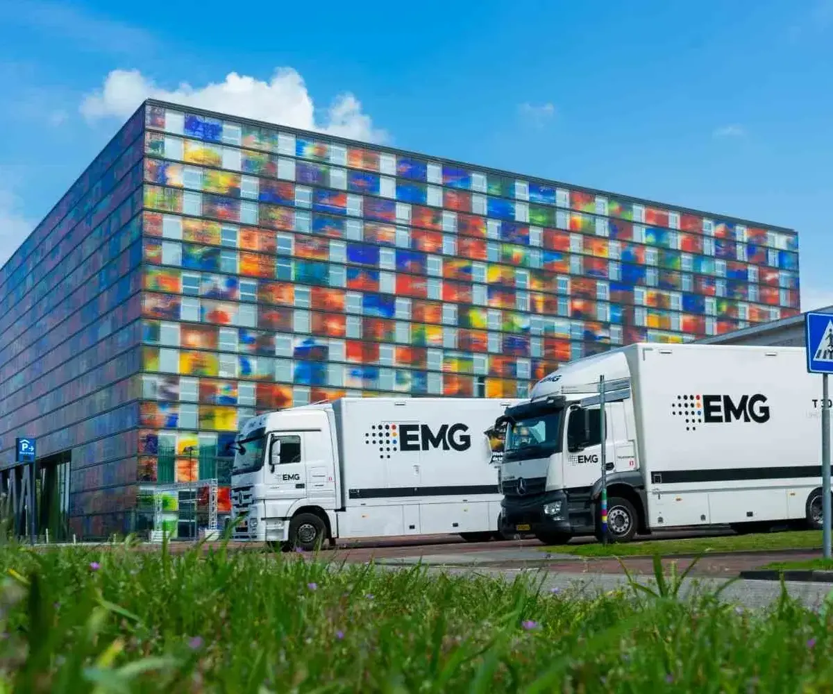 Two white EMG trucks parked outside a modern colorful glass building.