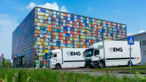 Two white EMG trucks parked outside a modern colorful glass building.