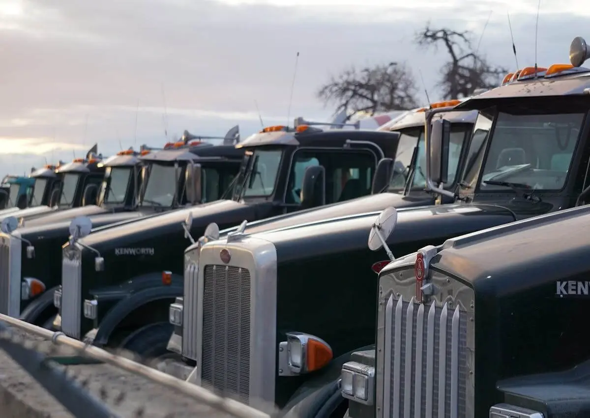 A row of black Kenworth semi-trucks parked side by side in a lot under a cloudy sky.
