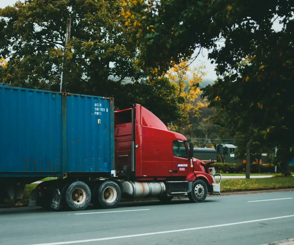 Red semi-truck hauling blue container on roadside.