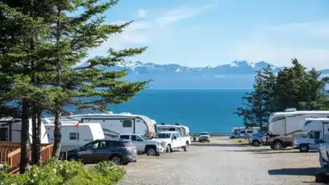 RVs and cars parked at a campground by the water with mountains in the background.