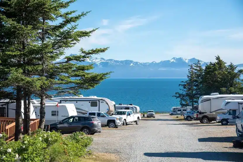 RVs and cars parked at a campground by the water with mountains in the background.