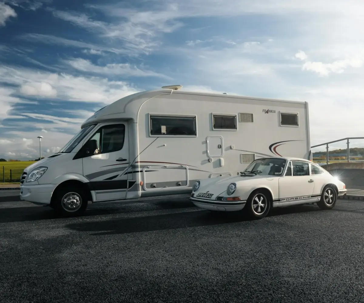RV and classic Porsche parked side-by-side on smooth asphalt under a dramatic sky.
