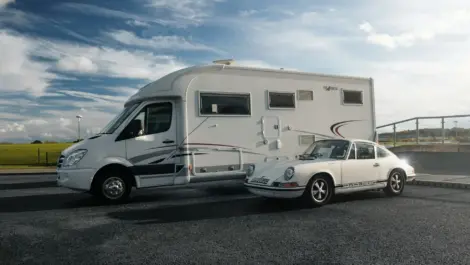 RV and classic Porsche parked side-by-side on smooth asphalt under a dramatic sky.
