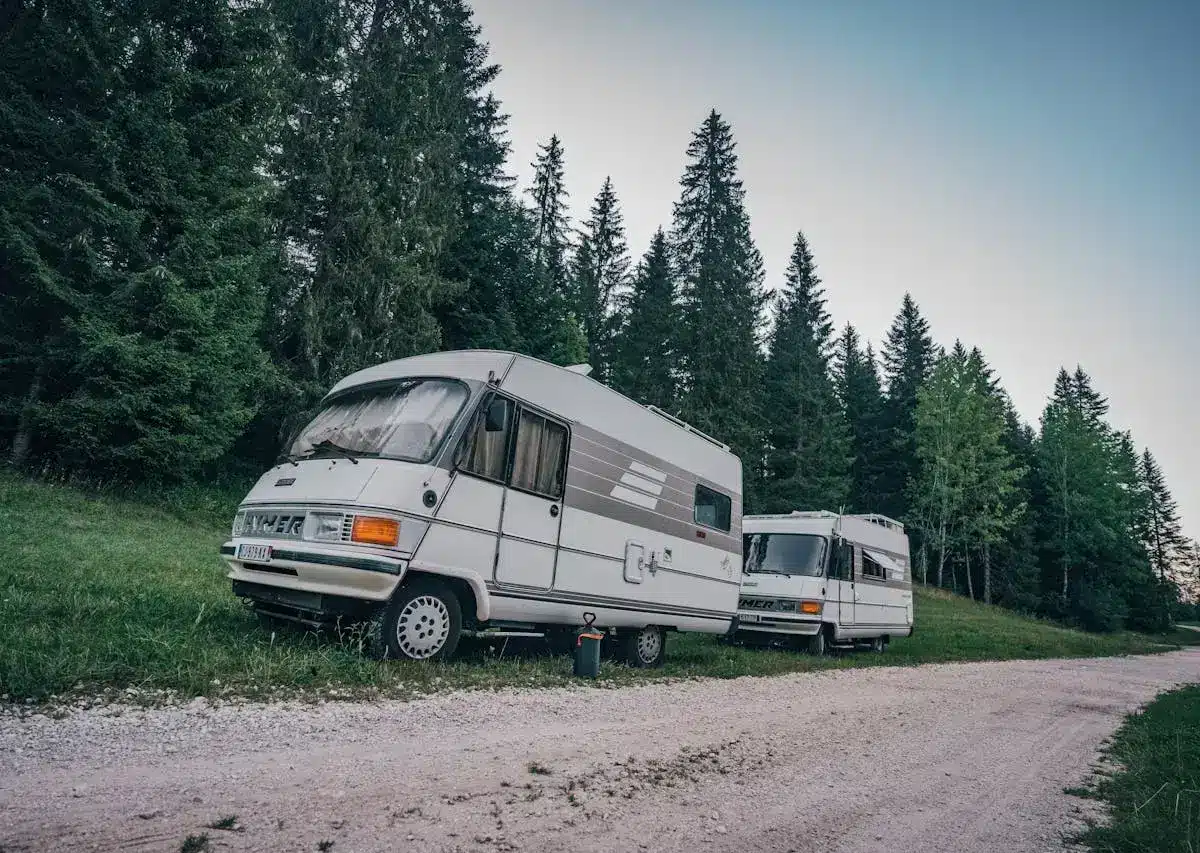 Two motorhomes parked on a gravel road near a forest.