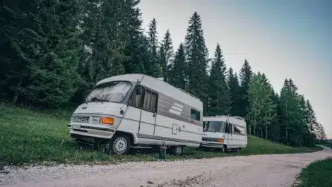 Two motorhomes parked on a gravel road near a forest.