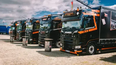 Row of black and orange Scania trucks parked outdoors under cloudy sky.