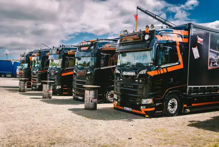 Row of black and orange Scania trucks parked outdoors under cloudy sky.