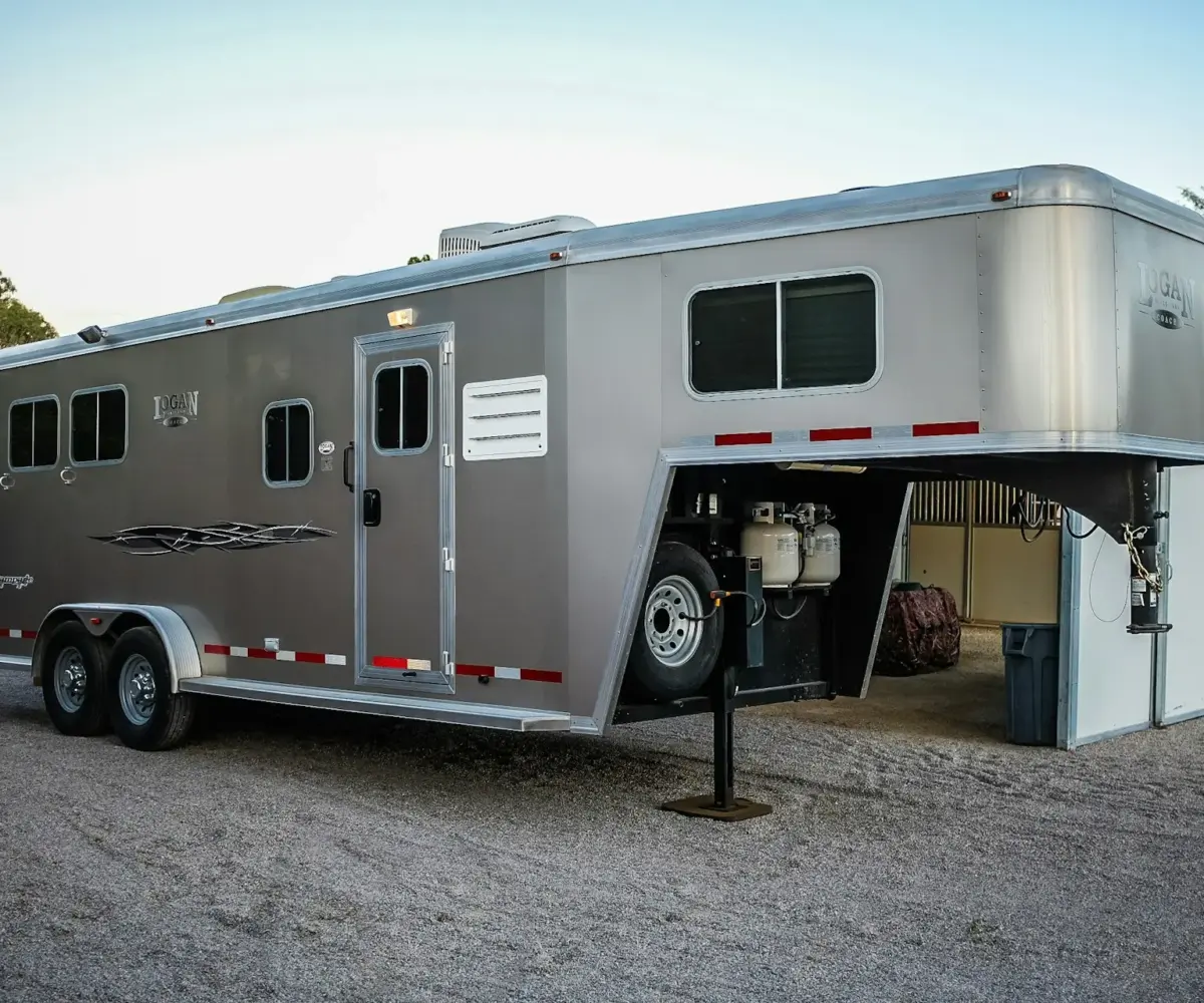 Silver gooseneck horse trailer parked on gravel beside a barn.