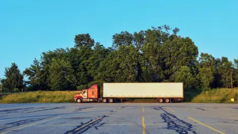 Red semi-truck with a white trailer parked alone in an empty lot.