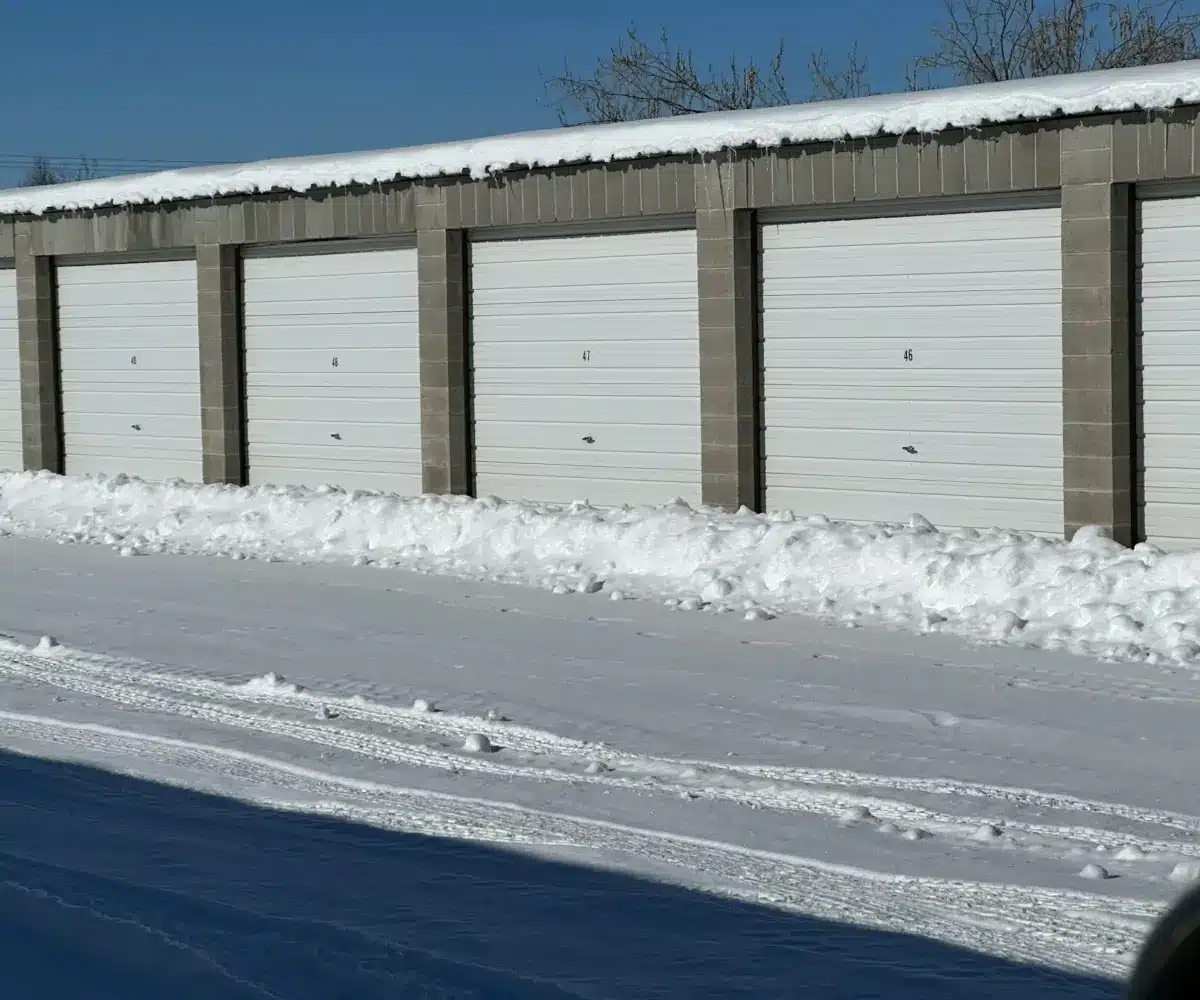 Row of snow-covered outdoor storage units with roll-up doors on a clear winter day.