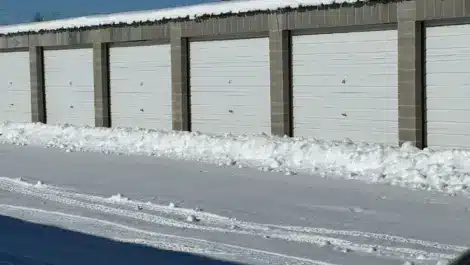 Row of snow-covered outdoor storage units with roll-up doors on a clear winter day.