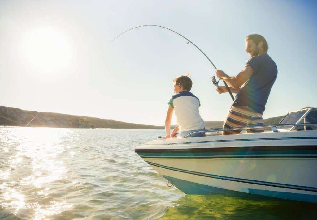 A father and son stand on a boat while the dad fishes in open water.