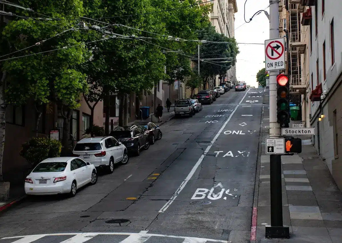Urban street with cars parked along the curb, traffic signals, and road markings indicating parking restrictions and city congestion.