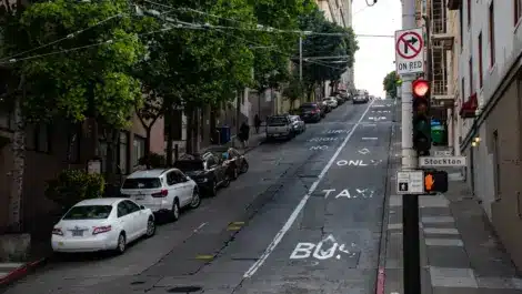 Urban street with cars parked along the curb, traffic signals, and road markings indicating parking restrictions and city congestion.