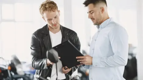 Two men reviewing motorcycle storage or insurance documents beside a bike, discussing coverage and protection options.